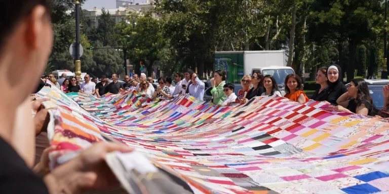 A large group of diverse people holding a massive colorful patchwork quilt outdoors