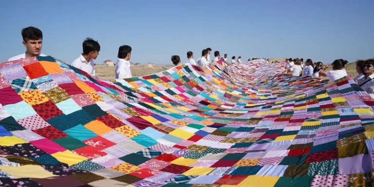 A large multicolored patchwork quilt is held by a group of people outdoors on a sunny day